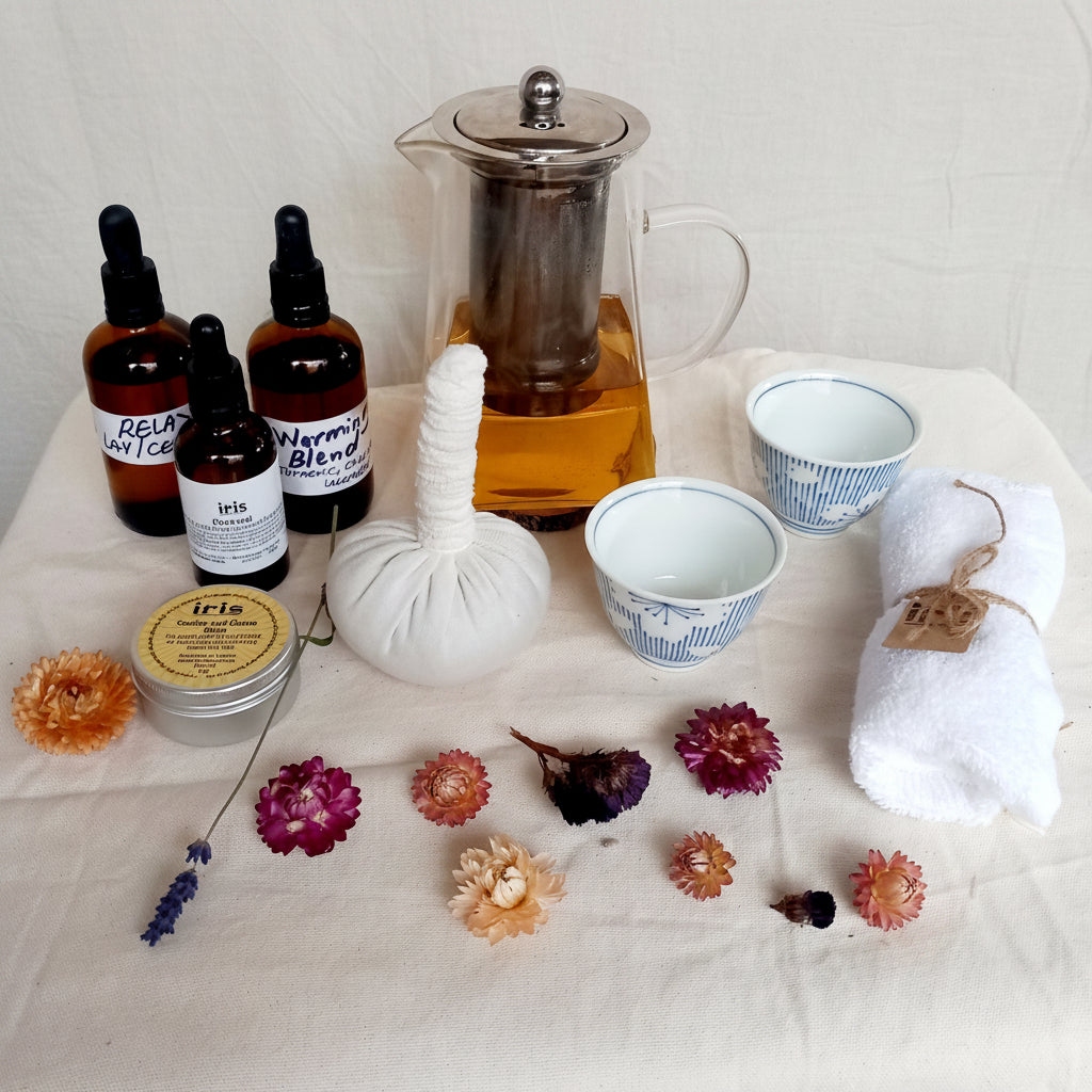 A tea preparation and brewing set arranged on a white fabric surface, including amber glass dropper bottles, a clear glass teapot with amber tea, two blue-and-white ceramic cups, a white wrapped hot water bag, a rolled white towel tied with twine, dried flowers, and a small tin of tea blends.